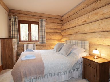 A bedroom with a double bed two blue and white towels on the bed wooden chest of drawers window with plaid curtains and a nightstand with a lamp at Moorhen Lodge in North Kessock