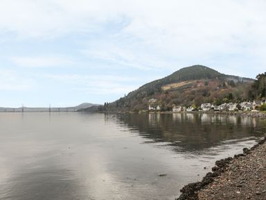 A shoreline with pebbles and seaweed beside a calm body of water with houses and a tree-covered hill in the background at Moorhen Lodge in North Kessock