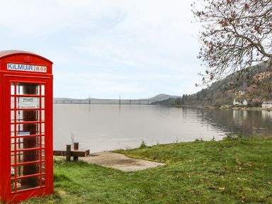 A red phone box near a grassy shore with a body of water and a bridge in the background at Moorhen Lodge in North Kessock
