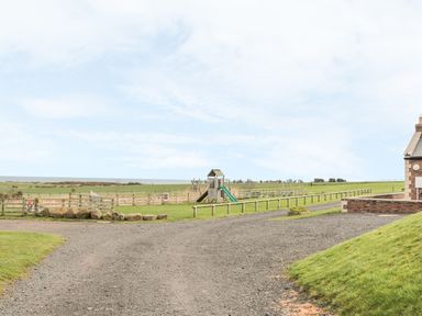 A playground with a slide and swings near a gravel path and grassy fields at St. Cruz in Embleton