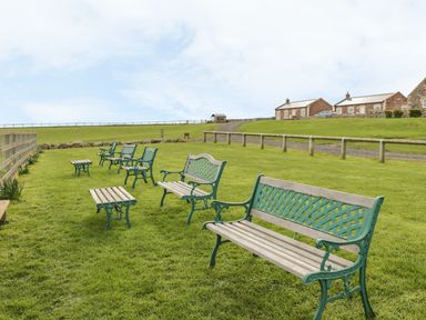 A grassy outdoor area with several benches and small tables near a wooden fence and stone buildings at St  Cruz in Embleton