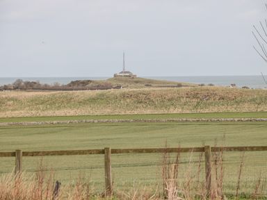 A grassy field with a wooden fence in front and a building with an antenna on a hill near the sea at St Cruz in Embleton