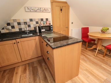 A kitchen with wooden cabinets a black countertop a sink stovetop microwave kettle a round wooden dining table with red chairs and fruit bowl at St Cruz in Embleton