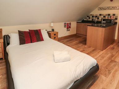 A bedroom with a double bed with white linens and red and brown pillows next to wooden nightstands and a kitchenette with a sink microwave and tiled backsplash at St Cruz in Embleton