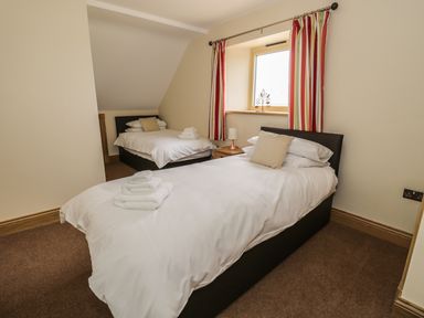 A bedroom with two single beds white bedding striped curtains and a small table with a lamp at Newton View Embleton