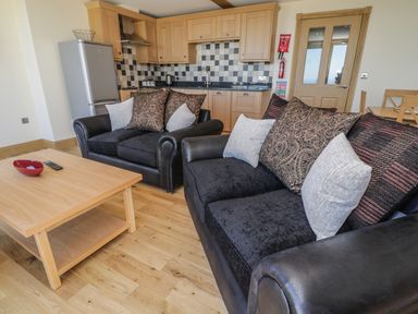 A living area with two black sofas with cushions and a wooden coffee table in front of a kitchen with wooden cabinets and tiled backsplash at Newton View in Embleton