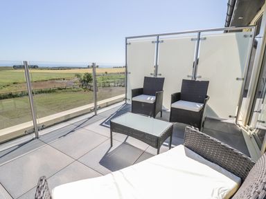 A balcony with two chairs a glass table and a cushioned sun lounger overlooking fields at Newton View in Embleton