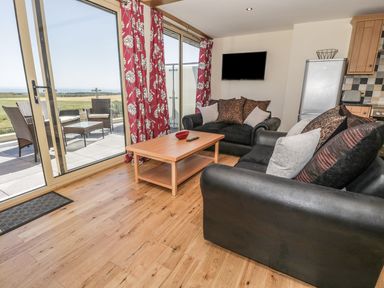 A living room with black sofas and cushions a wooden coffee table a wall mounted tv and glass doors leading to a patio with outdoor furniture at Newton View in Embleton