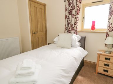 A bedroom with a single bed white bedding and towels on it a wooden door a window with red and white curtains and a bedside table with a lamp at Mill View in Embleton
