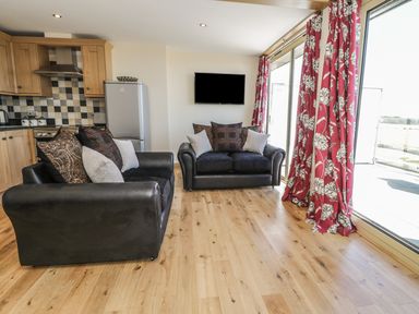 An open living area with two black sofas with cushions a wall-mounted television wooden kitchen cabinets and large sliding glass doors with red floral curtains at Mill View in Embleton