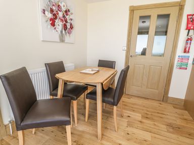 A small dining area with a wooden table and four brown chairs a wooden door and a floral painting on the wall at Mill View in Embleton