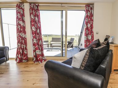 A living room with black leather sofas red floral curtains and glass doors leading to a patio with outdoor furniture at Mill View in Embleton