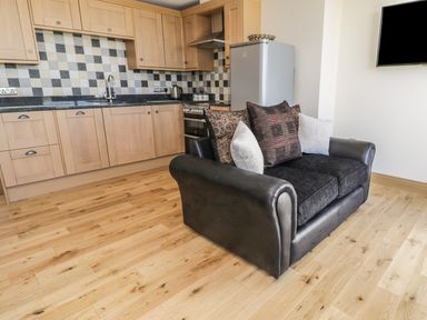 A kitchen with wooden cabinets a checkered tile backsplash a black sofa with cushions and a wall-mounted television at Mill View in Embleton
