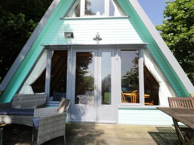 The exterior of an A-frame house with glass doors and windows showing a wooden dining table and chairs inside a patio with outdoor wicker furniture and a wooden table at Apple Tree Lodge in Gillingham