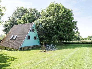 A triangular cabin with blue siding next to a patio set and wood stove in a grassy yard with trees at Apple Tree Lodge in Gillingham