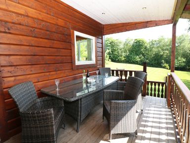 A wooden balcony with a glass table and wicker chairs overlooking a grassy area at Kipling Lodge in Rudyard