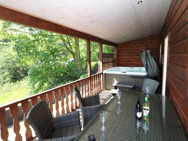 A covered outdoor balcony with a wicker table and chairs and a hot tub surrounded by wooden railing and trees at Kipling Lodge in Rudyard