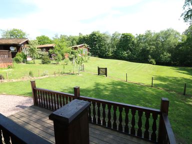 A wooden deck overlooking a fenced grassy yard with small trees and cabins in the background at Kipling Lodge in Rudyard
