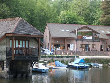 A riverside scene with boats docked near a boathouse and buildings with people outside at Kipling Lodge in Rudyard