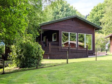 A wooden cabin with large windows and a porch surrounded by trees and grass at Kipling Lodge in Rudyard