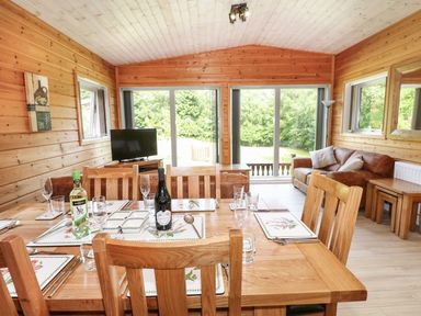 A wooden dining table with chairs bottles glasses and placemats in a wooden lounge with sofas a television and large windows at Kipling Lodge in Rudyard