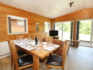 A dining area with a wooden table set for six and a living space with a TV and brown sofa at Kipling Lodge in Rudyard