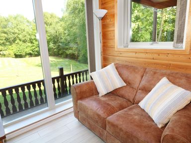 A living room with a brown sofa with pillows next to large windows showing a grassy yard at Kipling Lodge in Rudyard