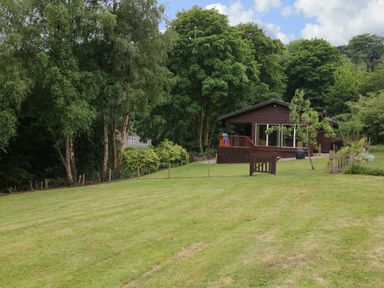 A wooden house with sliding glass doors surrounded by trees and a large grassy lawn at Kipling Lodge in Rudyard