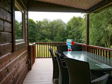 A covered wooden balcony with a glass table and wicker chairs overlooking trees and grass at Kipling Lodge in Rudyard