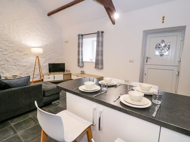 A kitchen and living area with a dining counter set for four people a gray sofa a TV on a wooden stand a floor lamp and a white door with a heart decoration at Cornel Glyd in Talybont near Barmouth