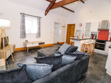 A living room with gray cushions on a sofa and a kitchen area with stools and a stove at Cornel Glyd in Talybont near Barmouth