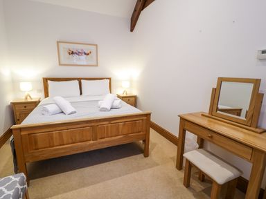 A bedroom with a wooden bed with white pillows and rolled towels beside wooden nightstands with lamps and a wooden dressing table with a mirror at Cornel Glyd in Talybont near Barmouth