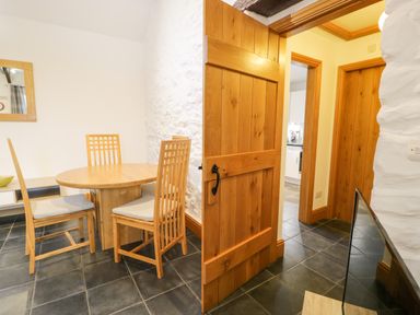 A dining area with a round wooden table and four wooden chairs near an open wooden door revealing a hallway at Sisial-y-Mor in Talybont near Barmouth