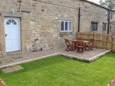 An outdoor patio area with a wooden table and chairs on gravel next to a stone building with a white door and a small fenced lawn at 1 Dunkirk Cottages Humshaugh near Hexham