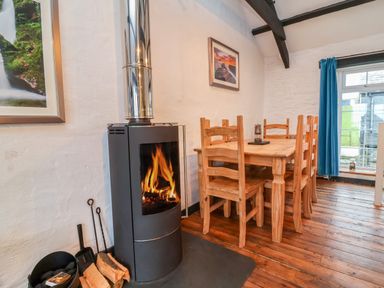 A wood burning stove with fire next to a wooden dining table and chairs in a room with wooden floor at Threshings Cottage in Tintagel