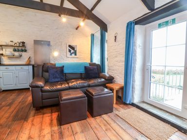 A living area with a brown leather sofa two ottomans wooden flooring and a kitchen area at Threshings Cottage in Tintagel