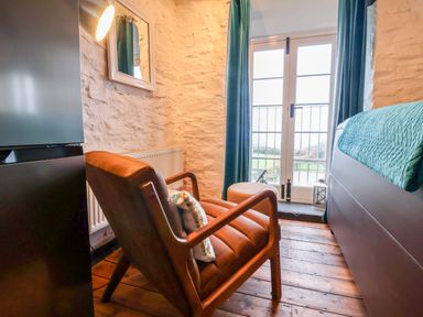 A small room with a brown armchair a side table a bed with a blue cover and glass doors with blue curtains at Threshings Cottage in Tintagel