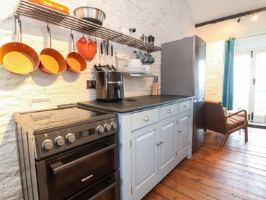 A kitchen with hanging pots and pans a stove counter with an air fryer dishes on shelves and a refrigerator with an armchair nearby at Threshings Cottage in Tintagel