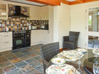 A kitchen with cream cabinets a black stove tiled backsplash and tiled floor beside a dining table set with plates glasses and wicker chairs at Fig Tree Cottage in Bickleigh