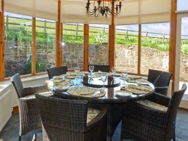 A dining area with a round glass table set with plates and glasses surrounded by wicker chairs in a room with large windows and a stone wall outside at Fig Tree Cottage in Bickleigh
