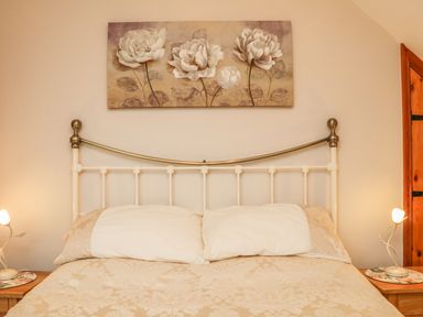 A bedroom with a bed with white pillows and beige bedding a floral painting above the bed and wooden bedside tables with lamps at Fig Tree Cottage in Bickleigh