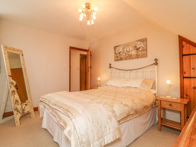A bedroom with a bed table lamps a standing mirror and floral artwork at Fig Tree Cottage in Bickleigh