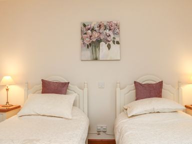 A bedroom with two single beds each with white bedding and a mauve pillow a floral painting on the wall and two bedside lamps at Fig Tree Cottage in Bickleigh