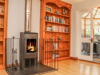 A wood burning stove with fire next to a wooden bookshelf with books and decorative items and a glass door leading to a dining area at Fig Tree Cottage in Bickleigh