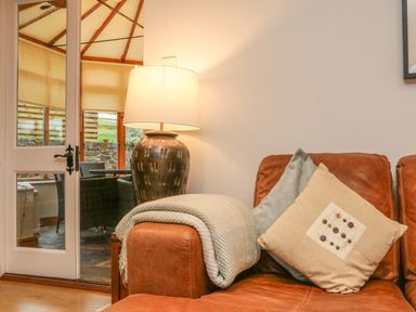 A brown leather sofa with two cushions and a blanket next to a ceramic lamp and glass door leading to a room with wicker chairs at Fig Tree Cottage in Bickleigh