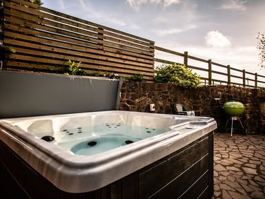 An outdoor hot tub on a stone patio with a wooden fence stone wall green barbecue and plastic chairs at Fig Tree Cottage in Bickleigh
