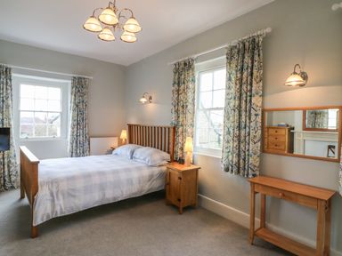 A bedroom with a wooden bed with side tables beside windows with patterned curtains and a wooden dressing table with a mirror at Tick Tock Cottage in Marnhull