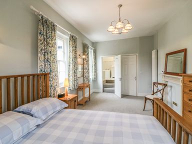 A bedroom with a wooden bed, bedside table with lamp, chair, dresser with mirror, curtains, and an open door at Tick Tock Cottage in Marnhull