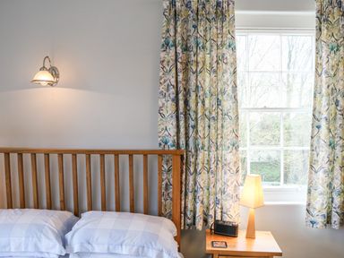 A bedroom with a wooden bed frame pillows patterned curtains a bedside table with a lamp and a radio at Tick Tock Cottage in Marnhull