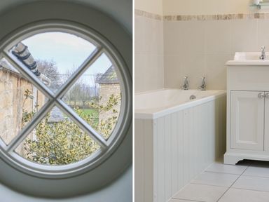 A round window with a view of stone buildings and trees and a bathroom with a bathtub and a sink unit at Tick Tock Cottage in Marnhull